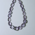 A beaded necklace made of smooth, oval-shaped translucent purple glass beads, arranged in a U-shape on a light gray background.