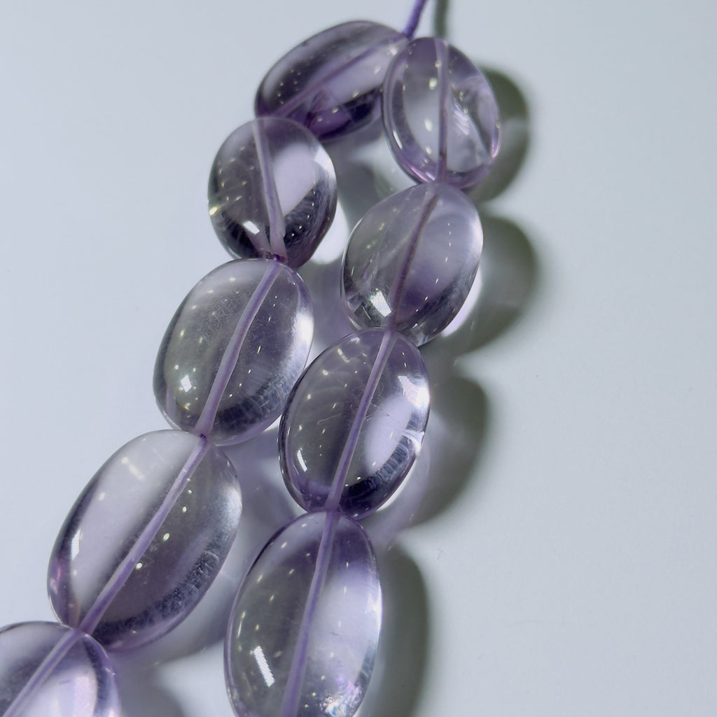 A close-up of translucent oval beads in a light purple or lavender tone, strung along a cord against a pale gray background.