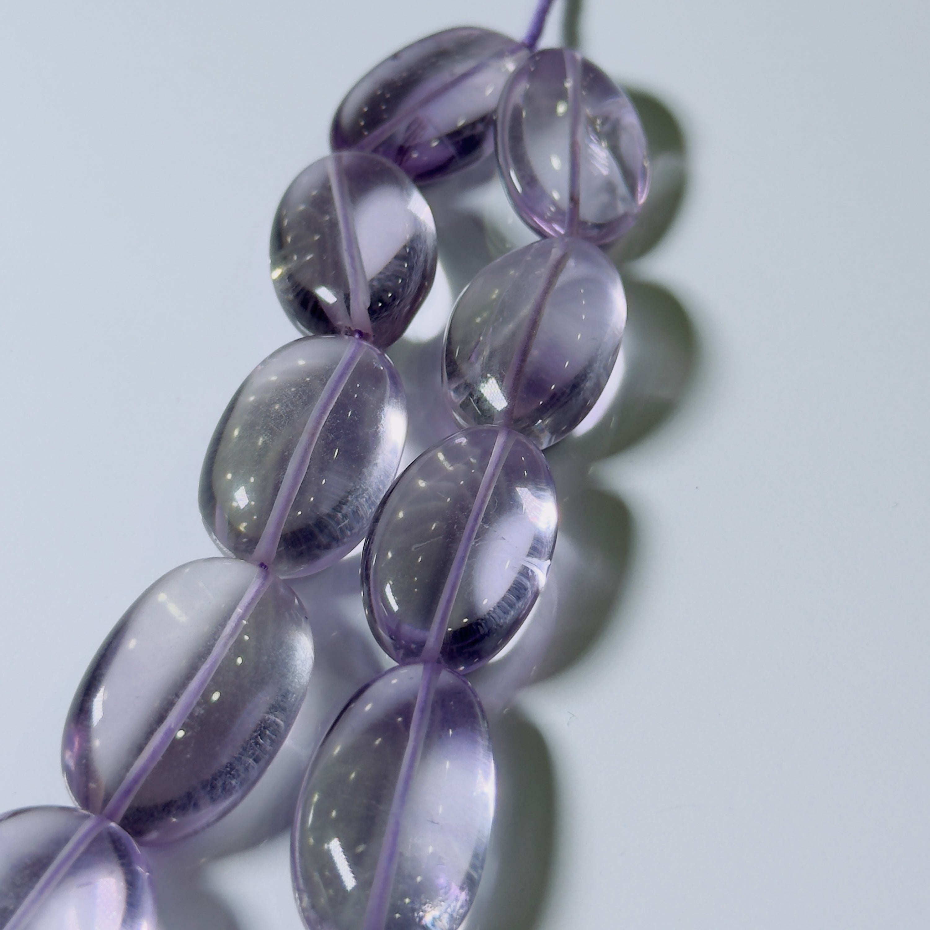 A close-up of translucent oval beads in a light purple or lavender tone, strung along a cord against a pale gray background.