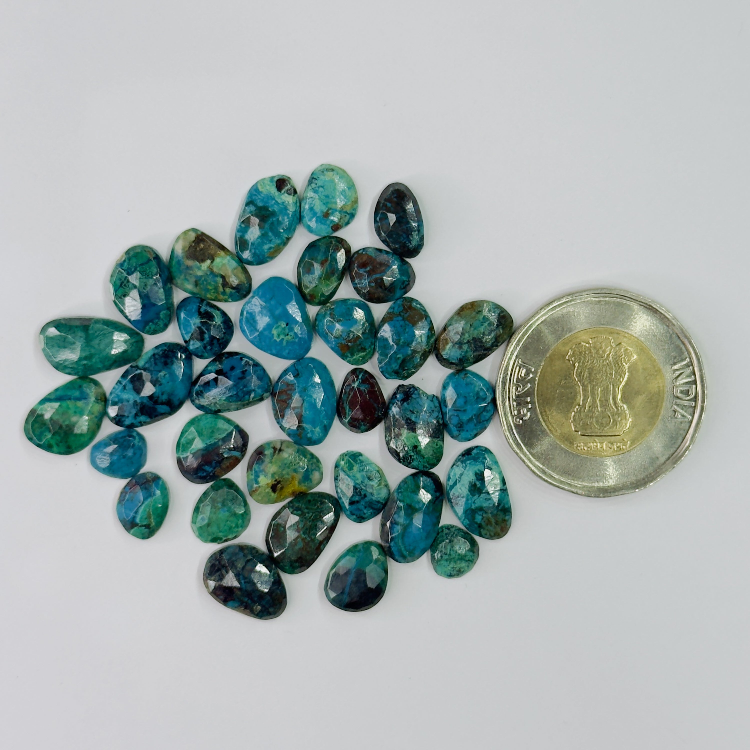 A group of polished tumbled gemstones in shades of teal, blue, and green, arranged next to a Indian 500 Rupee coin for scale, on a plain white background.