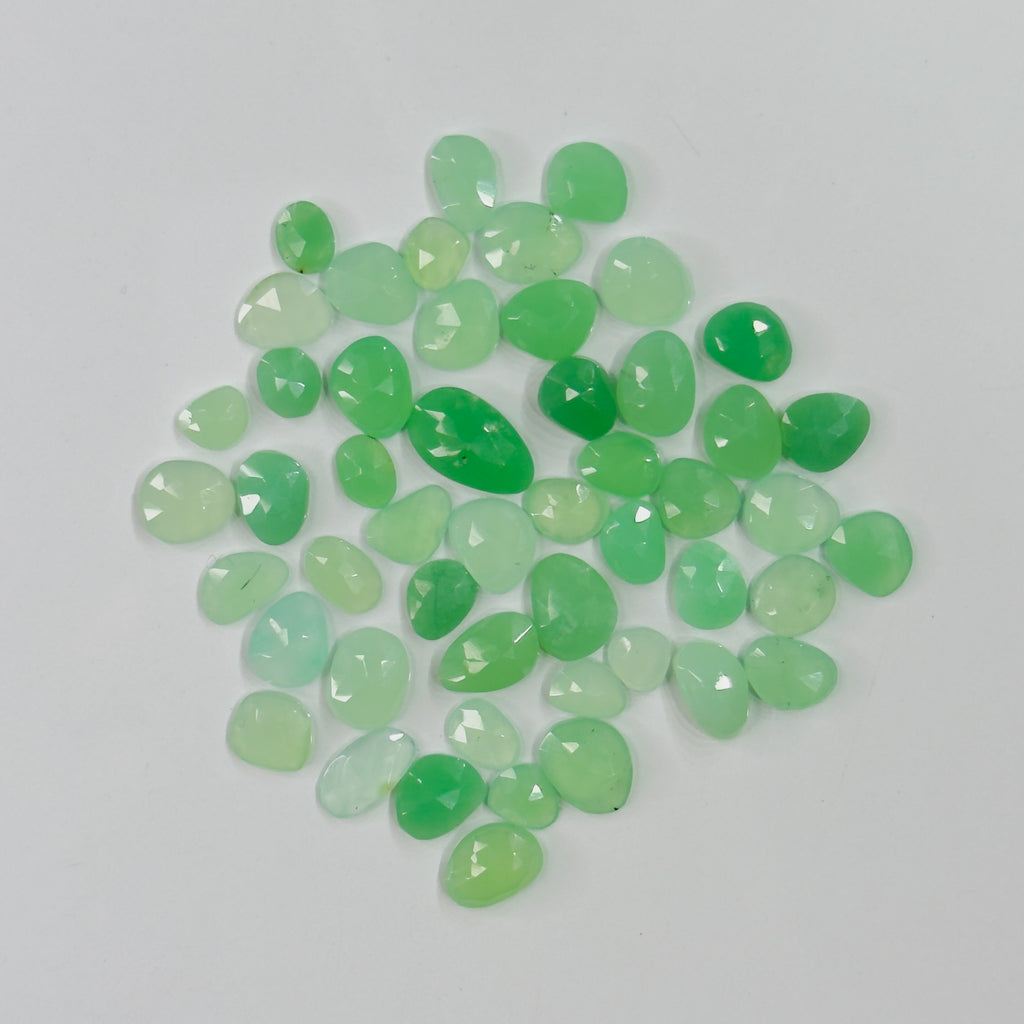 A pile of small, faceted green gemstones in various shades of light and medium green, arranged on a plain light gray background.