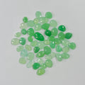 A pile of small, faceted green gemstones in various shades of light and medium green, arranged on a plain light gray background.