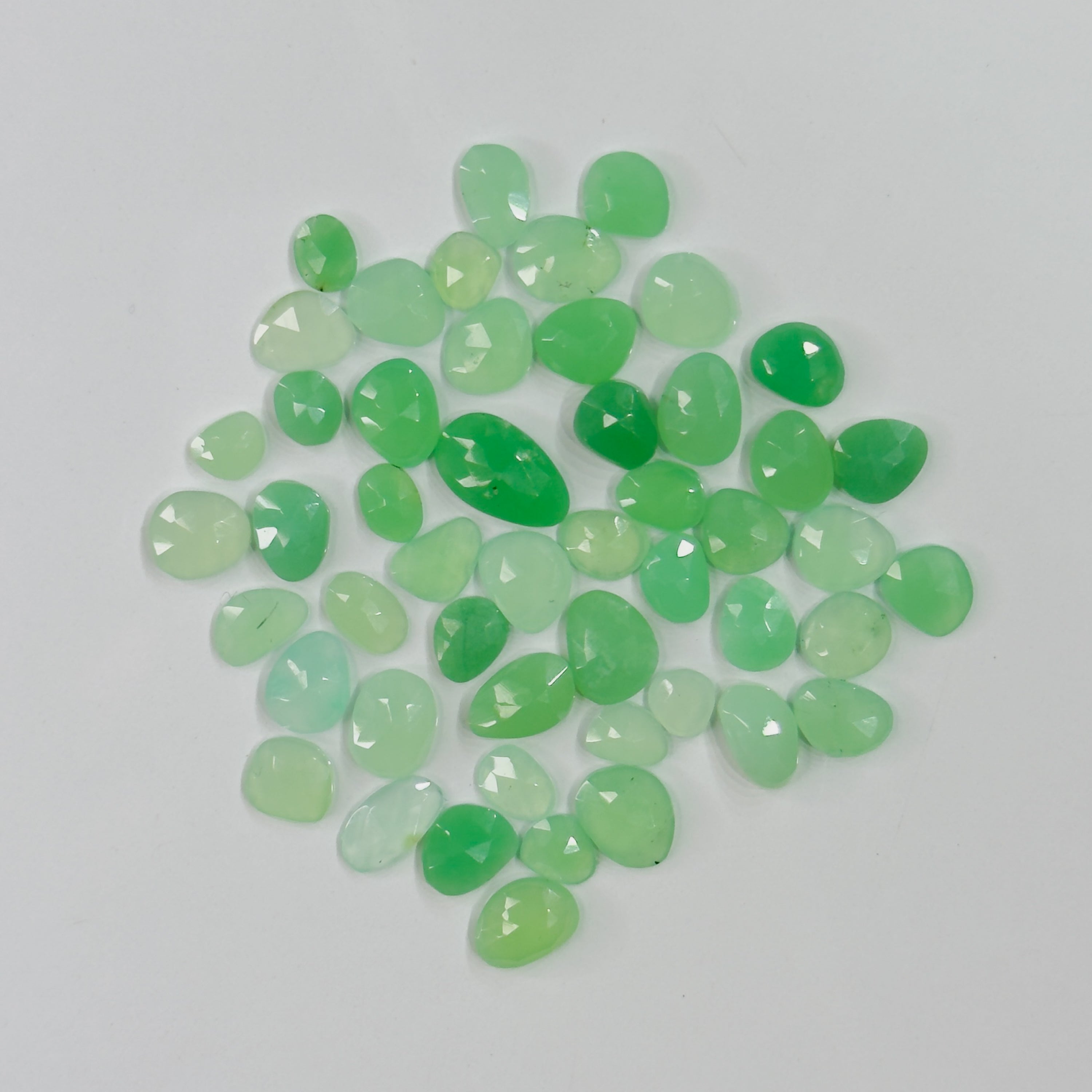 A pile of small, faceted green gemstones in various shades of light and medium green, arranged on a plain light gray background.