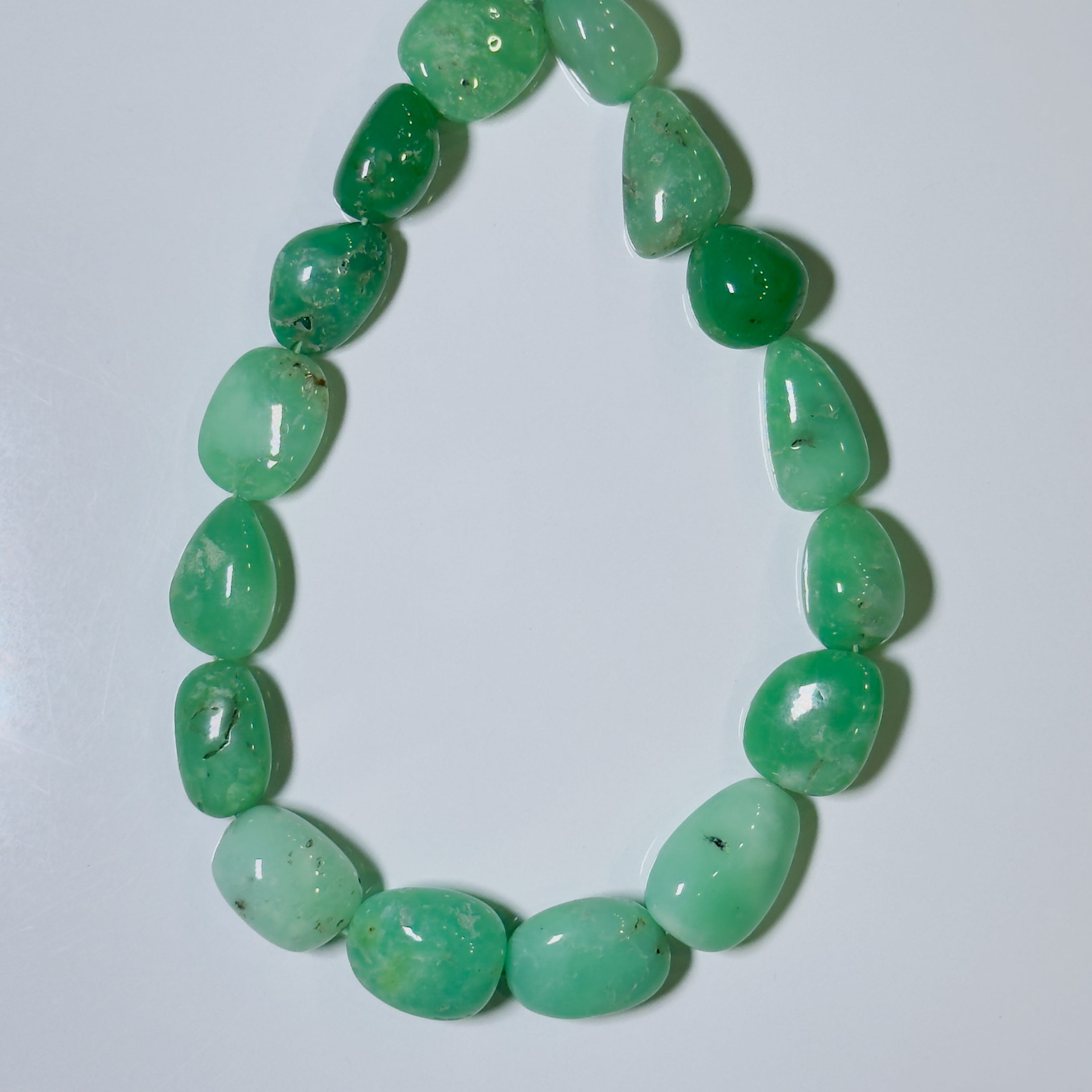 A beaded necklace made of polished, tumbled green gemstone beads with natural inclusions, arranged in an oval shape on a plain light gray background.