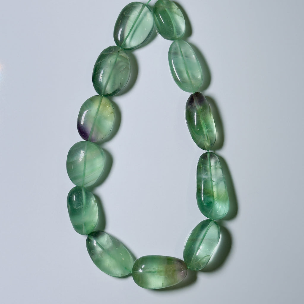 A beaded necklace made of polished green stone beads with natural inclusions and some purple undertones, arranged in a loop on a light gray background.