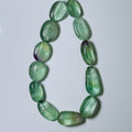 A beaded necklace made of polished green stone beads with natural inclusions and some purple undertones, arranged in a loop on a light gray background.