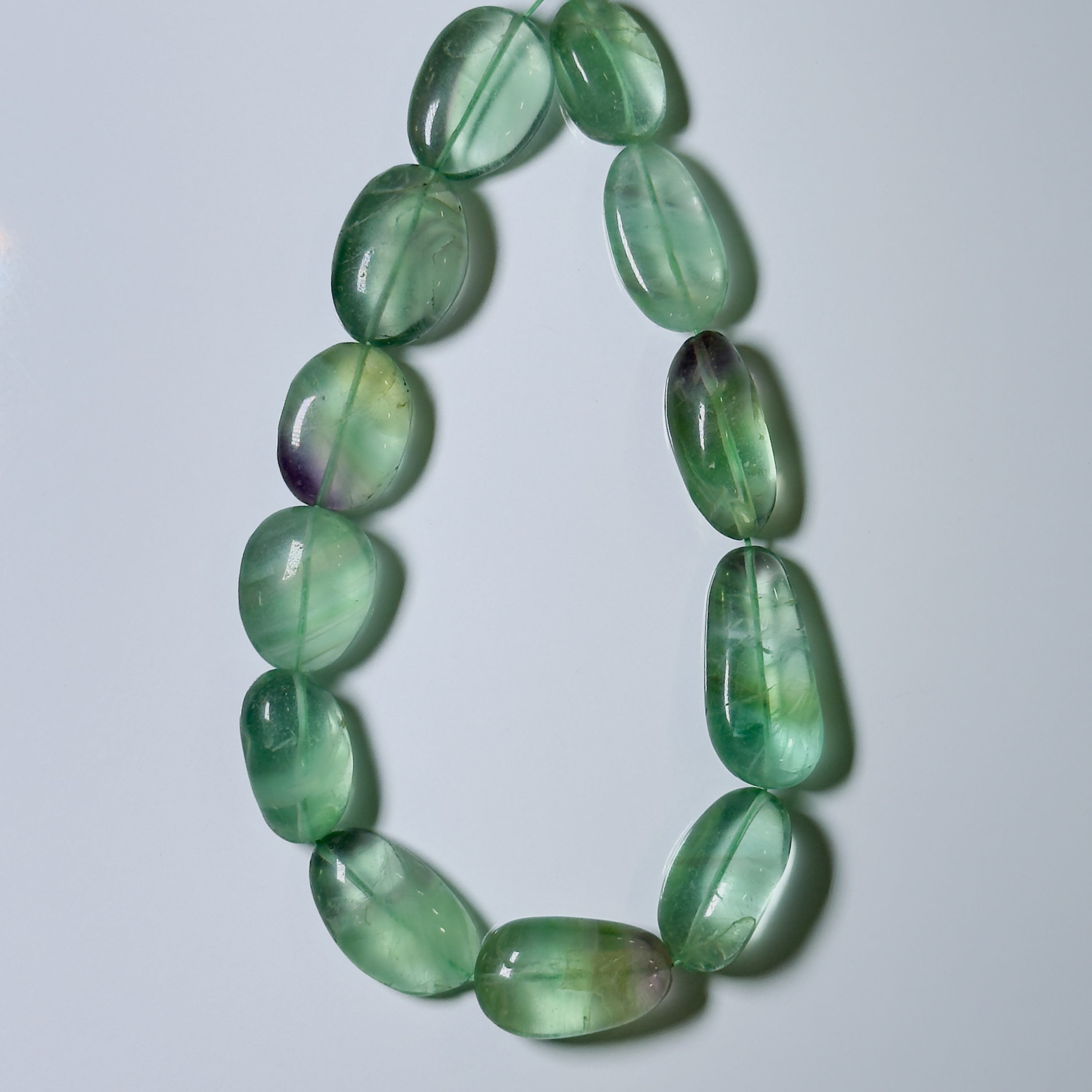 A beaded necklace made of polished green stone beads with natural inclusions and some purple undertones, arranged in a loop on a light gray background.
