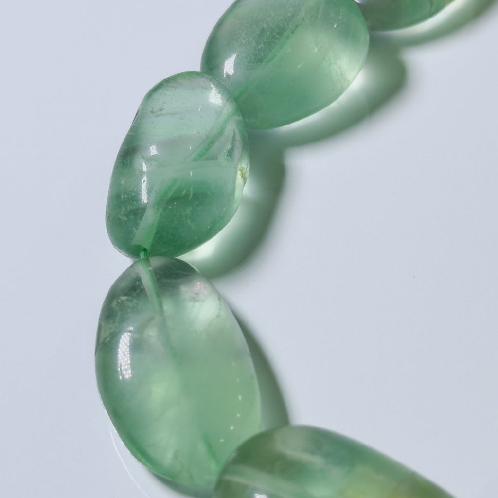 Close-up of a beaded strand with smooth, polished oval beads in a translucent light green stone, set against a clean light gray background.