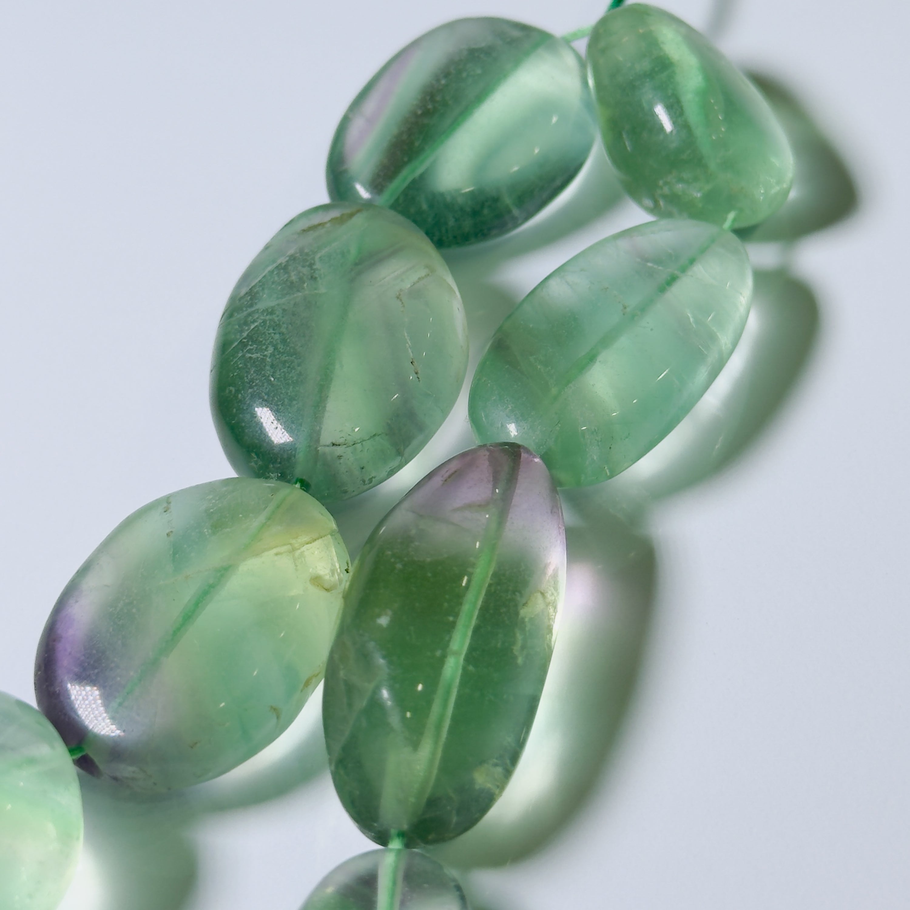A close-up of polished oval gemstone beads in a translucent light green color with subtle purple and blue iridescent tones, strung on a thin cord against a pale light gray background.