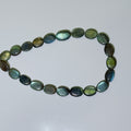 A beaded bracelet made of polished oval labradorite stones, showing a shimmering green and blue iridescent coloration, arranged in a loose circle on a light gray background.