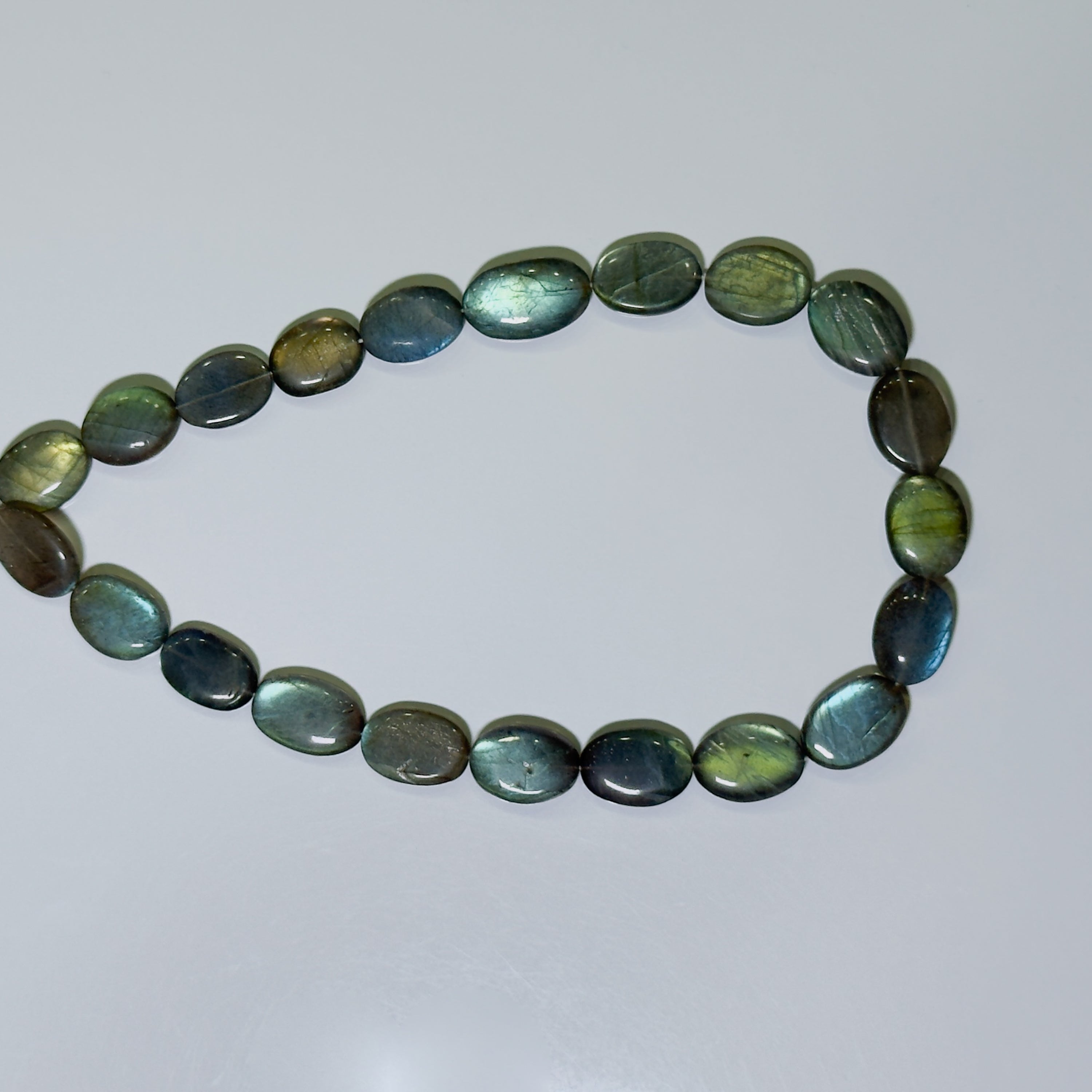 A beaded bracelet made of polished oval labradorite stones, showing a shimmering green and blue iridescent coloration, arranged in a loose circle on a light gray background.