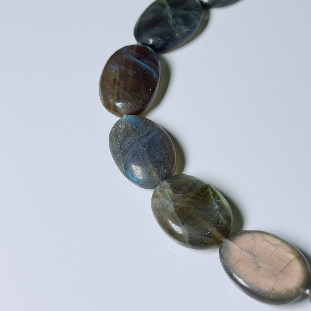 A close-up of polished oval labradorite gemstone beads arranged diagonally on a light gray background.