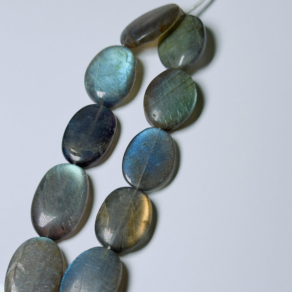 A strand of polished oval labradorite beads with a shimmering blue, green, and gray iridescent finish, laid diagonally on a light gray background.