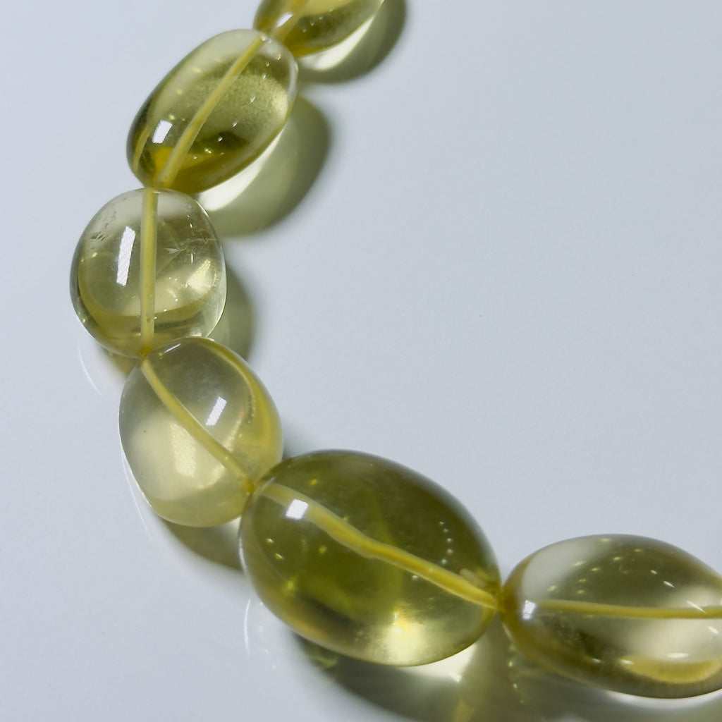 A close-up of a beaded strand with translucent oval beads in a pale yellow-green hue, arranged diagonally on a light gray reflective surface.