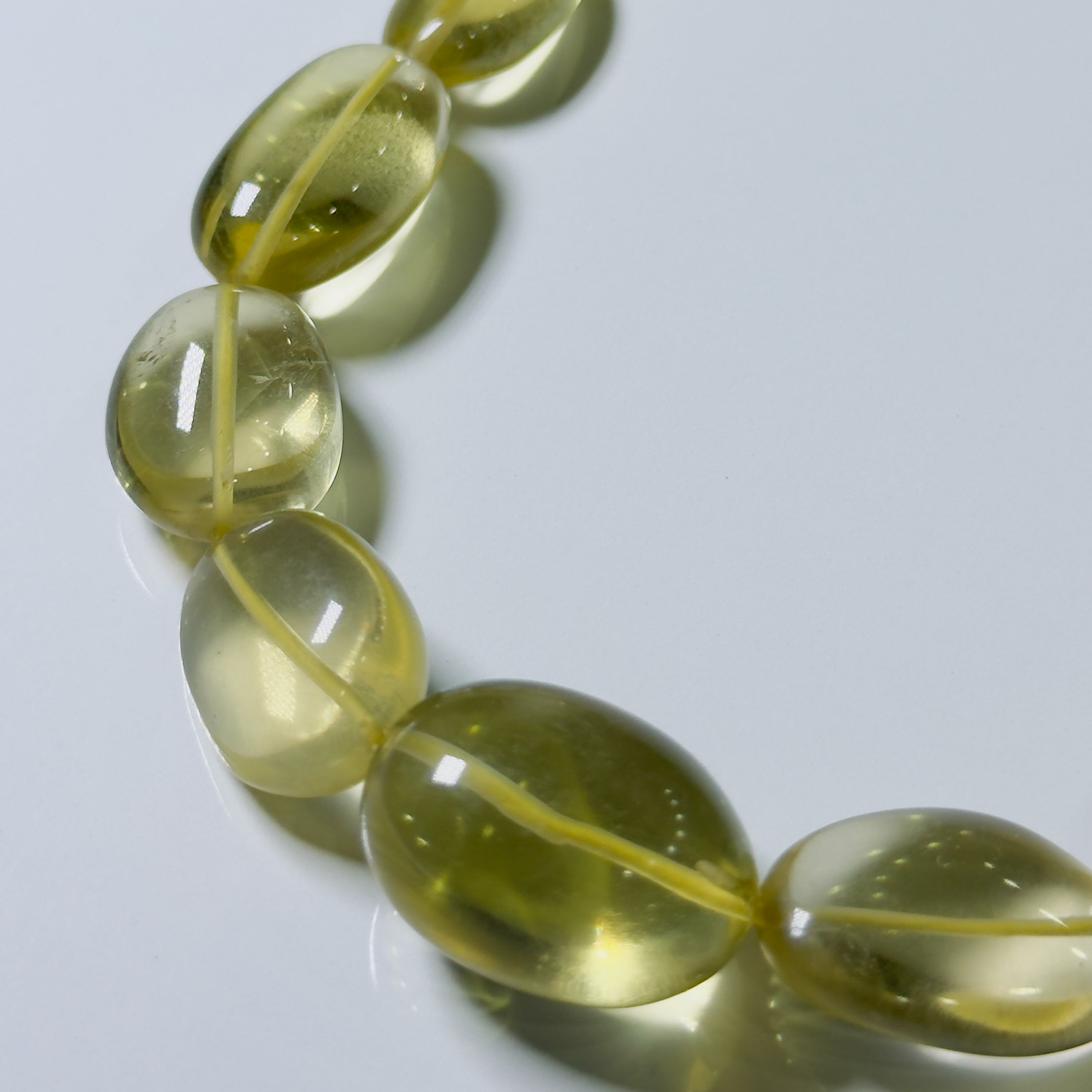A close-up of a beaded strand with translucent oval beads in a pale yellow-green hue, arranged diagonally on a light gray reflective surface.