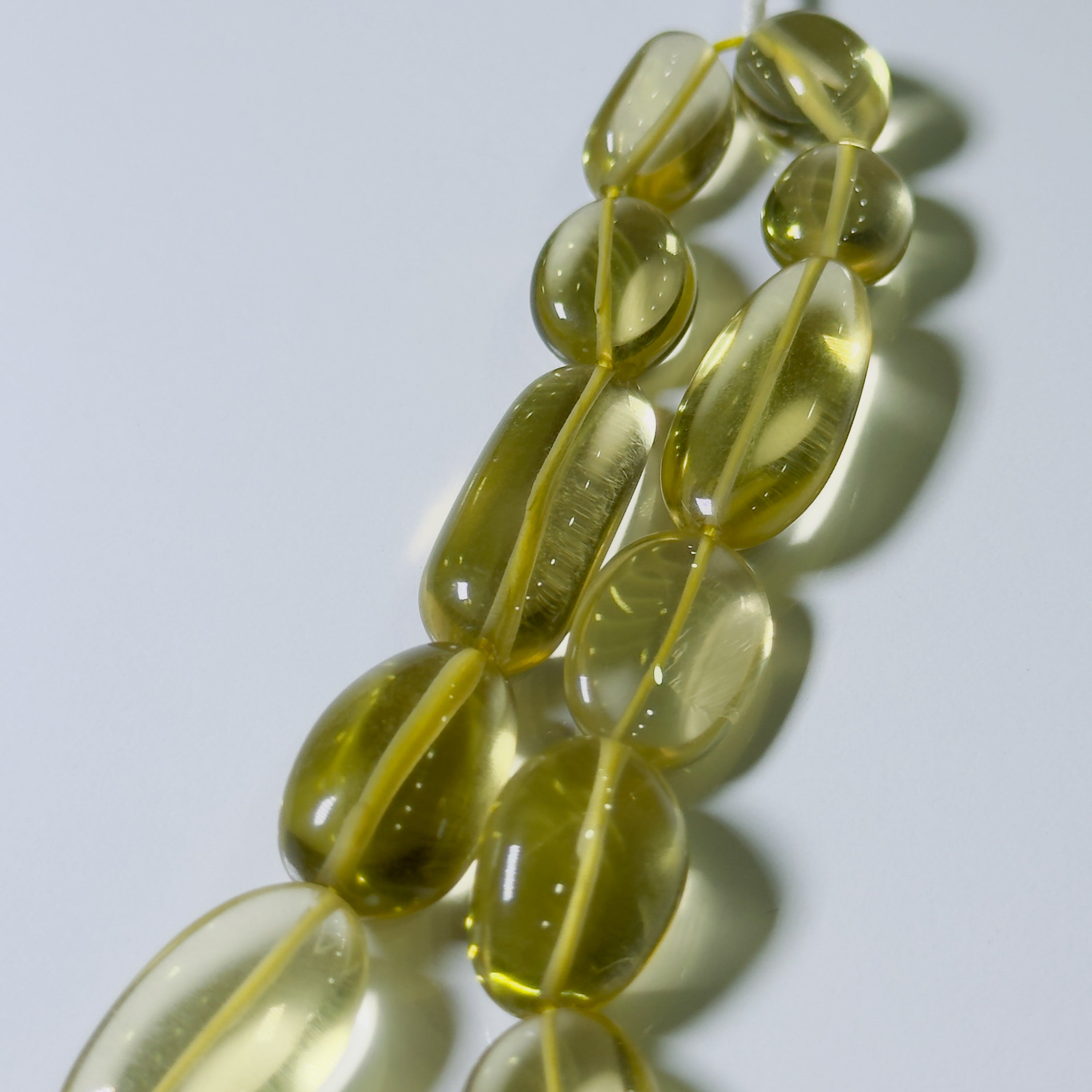A close-up of translucent olive-yellow glass beads strung on a cord, arranged diagonally against a light gray background with soft shadows.
