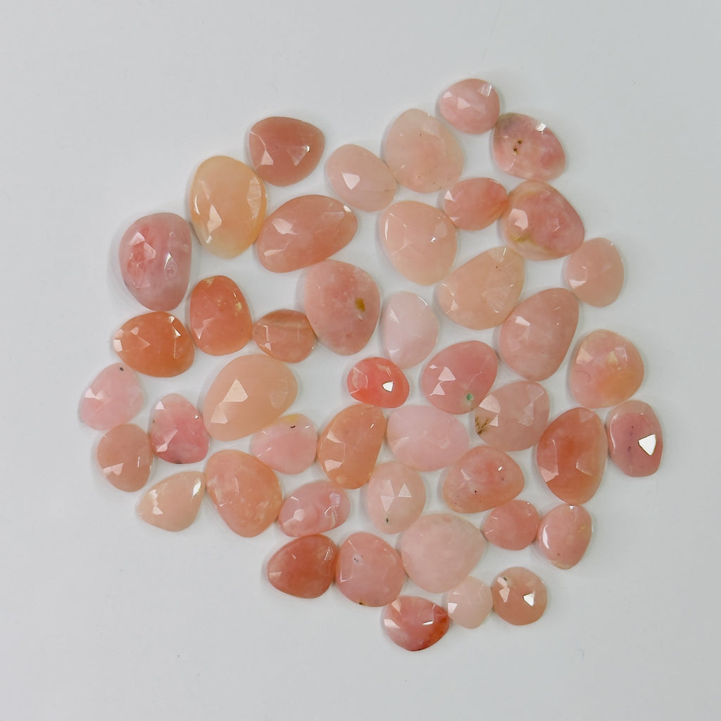 A pile of faceted, teardrop-shaped gemstone cabochons in shades of pink, peach, and light orange, arranged on a plain light gray background.