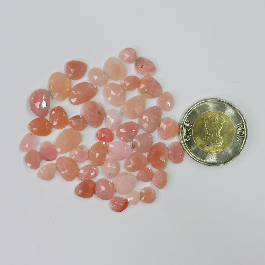 A pile of small faceted peach-pink gemstones next to a Indian 25-paise coin for size reference, on a plain light background.