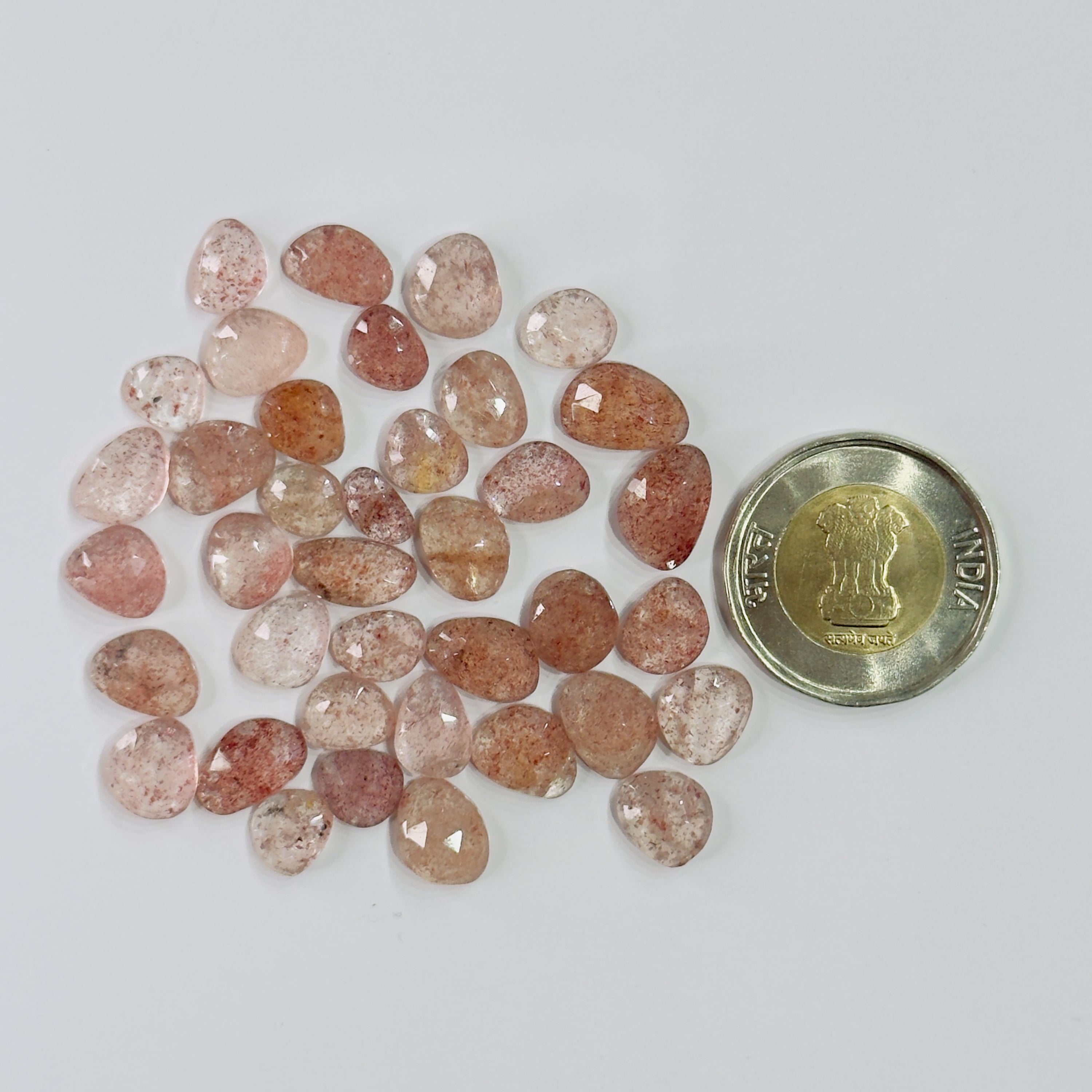 A pile of small polished peach-pink and translucent brown gemstones next to a gold-colored Indian 50-paise coin for scale, on a plain light gray background.