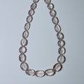 A beaded necklace made of translucent oval beads with a soft pinkish-gray hue, arranged in a teardrop shape against a light gray background.