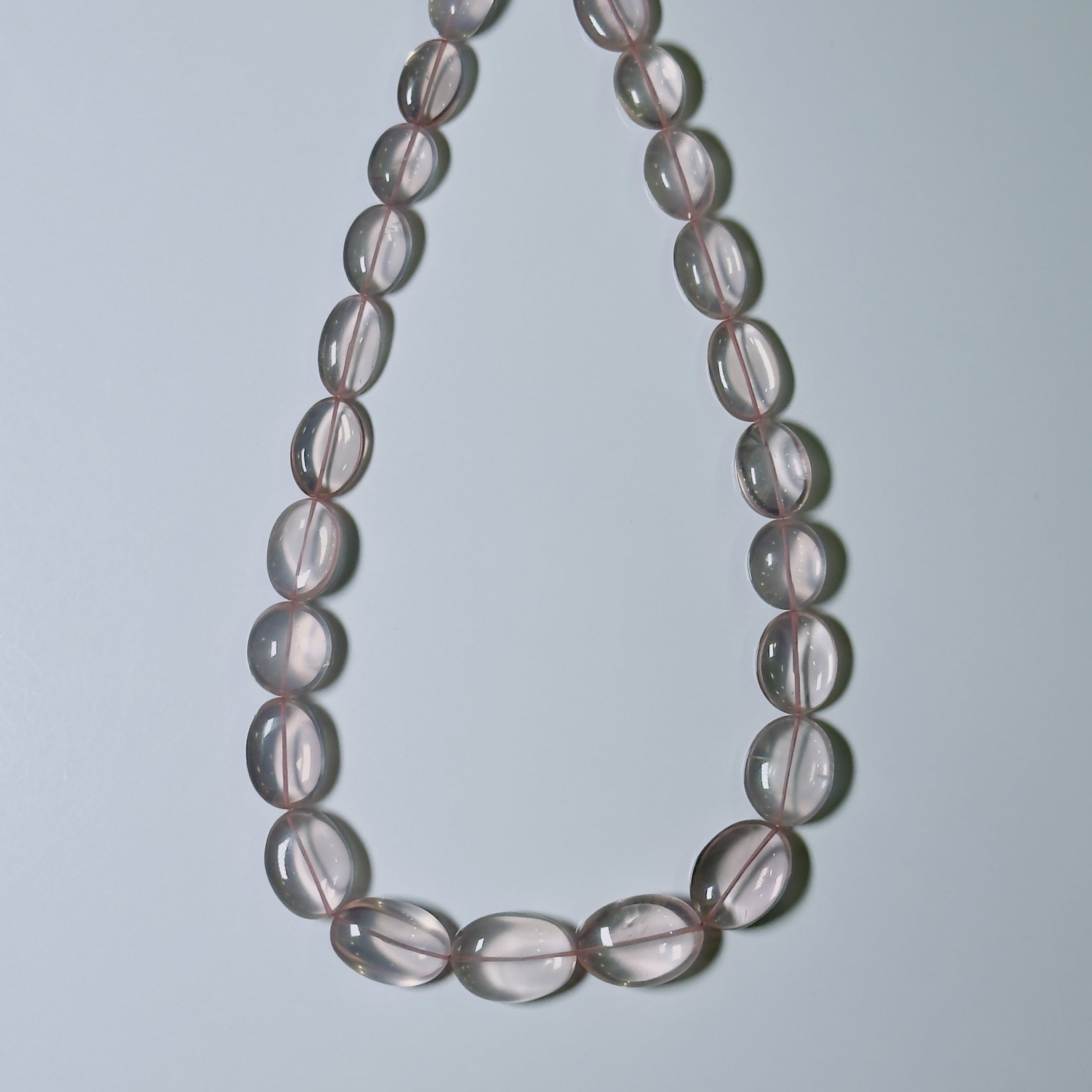 A beaded necklace made of translucent oval beads with a soft pinkish-gray hue, arranged in a teardrop shape against a light gray background.