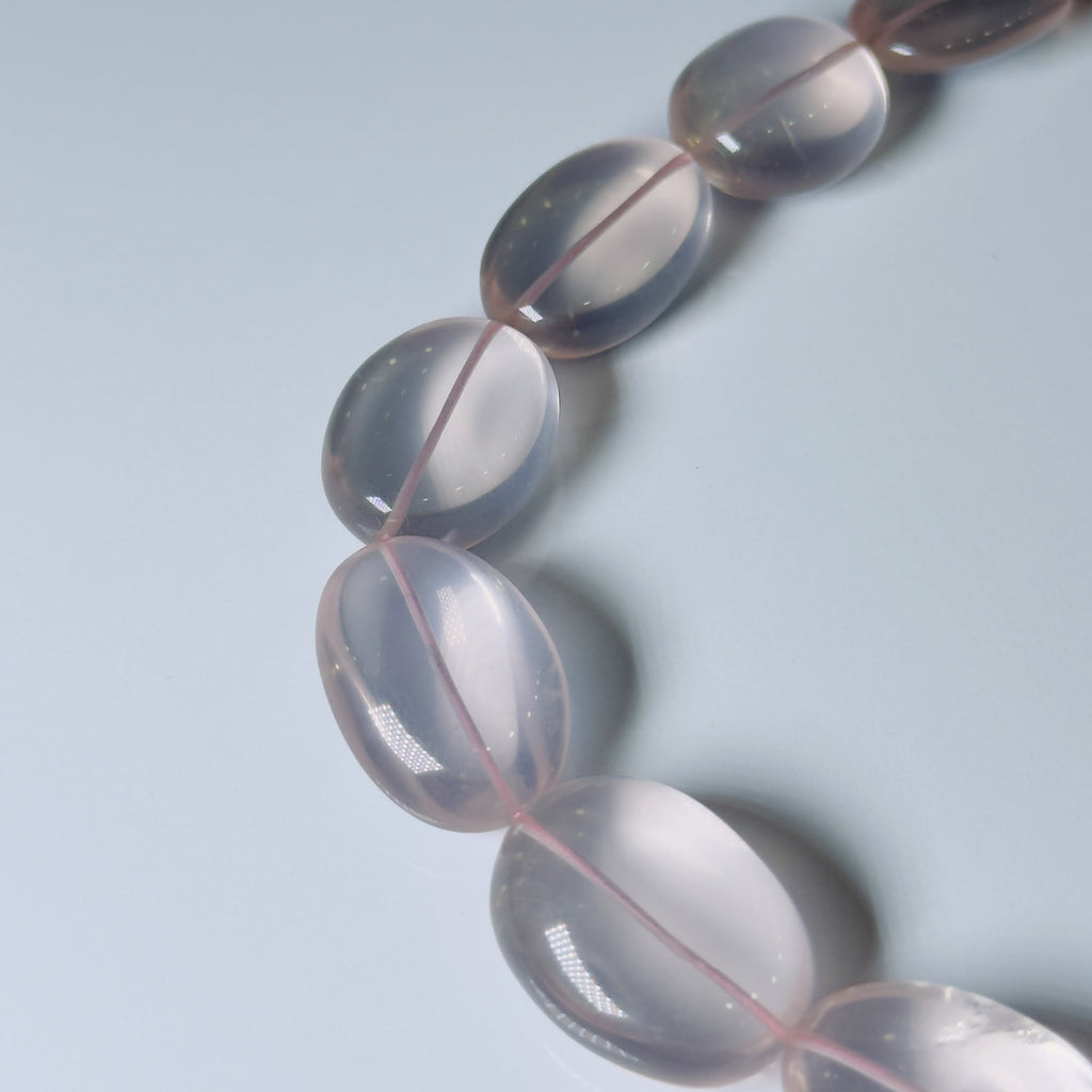 A close-up of a translucent pale pink beaded strand, likely a bracelet or necklace, with smooth oval beads on a light blue-gray background.