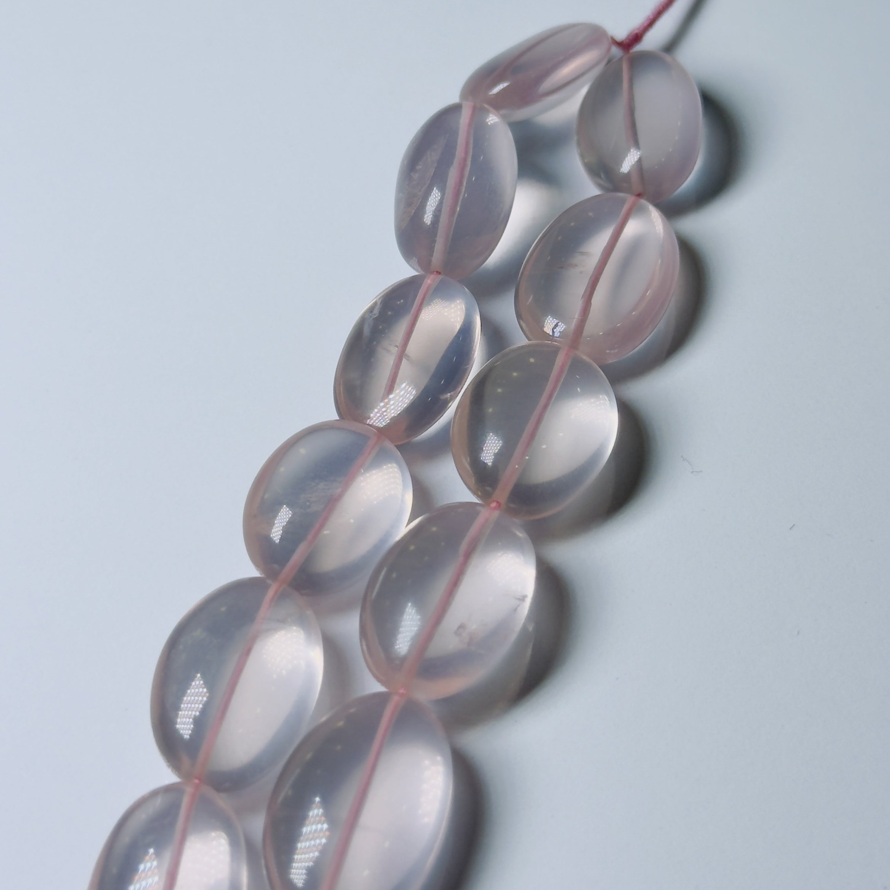 A strand of translucent pale pink oval beads strung on a pink thread, photographed on a light gray background.