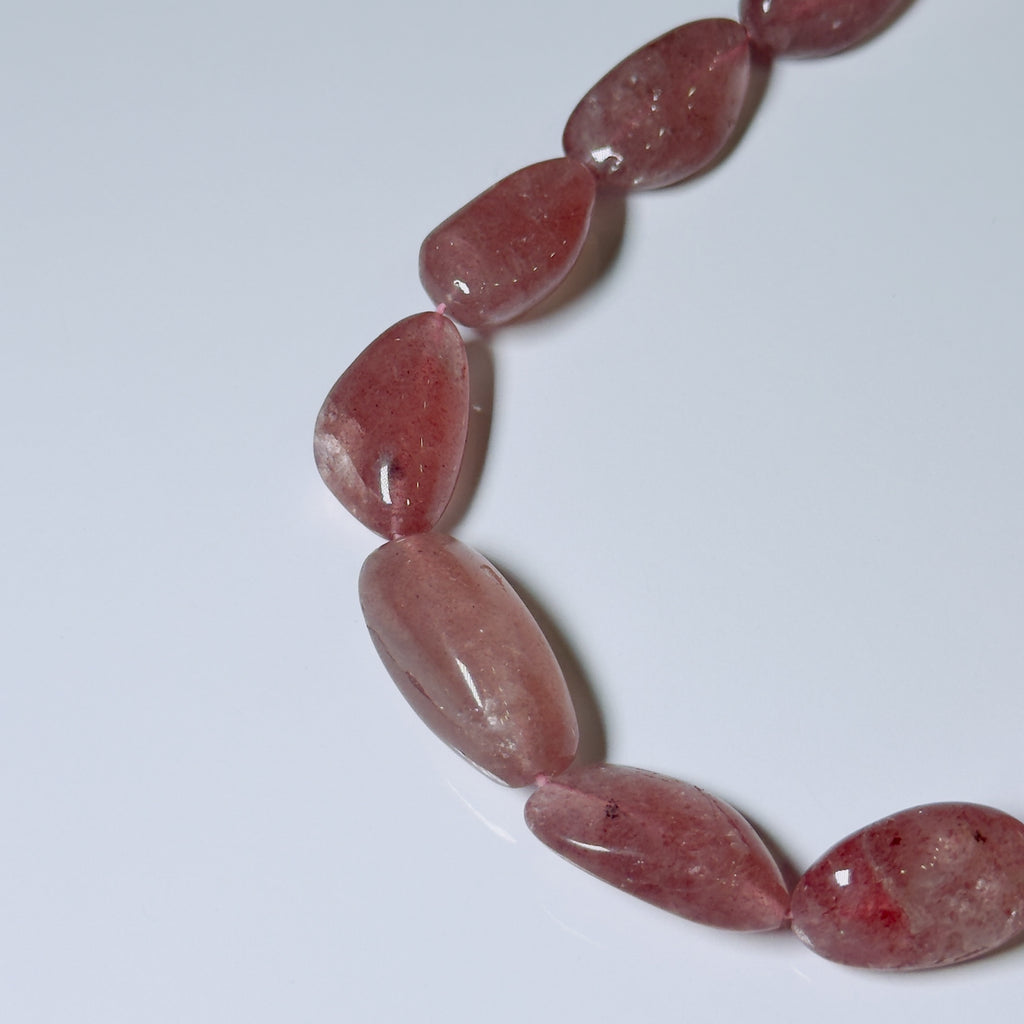 A close-up of polished teardrop-shaped beads in a dusty rose pink color, arranged in a curved line on a plain light gray background.