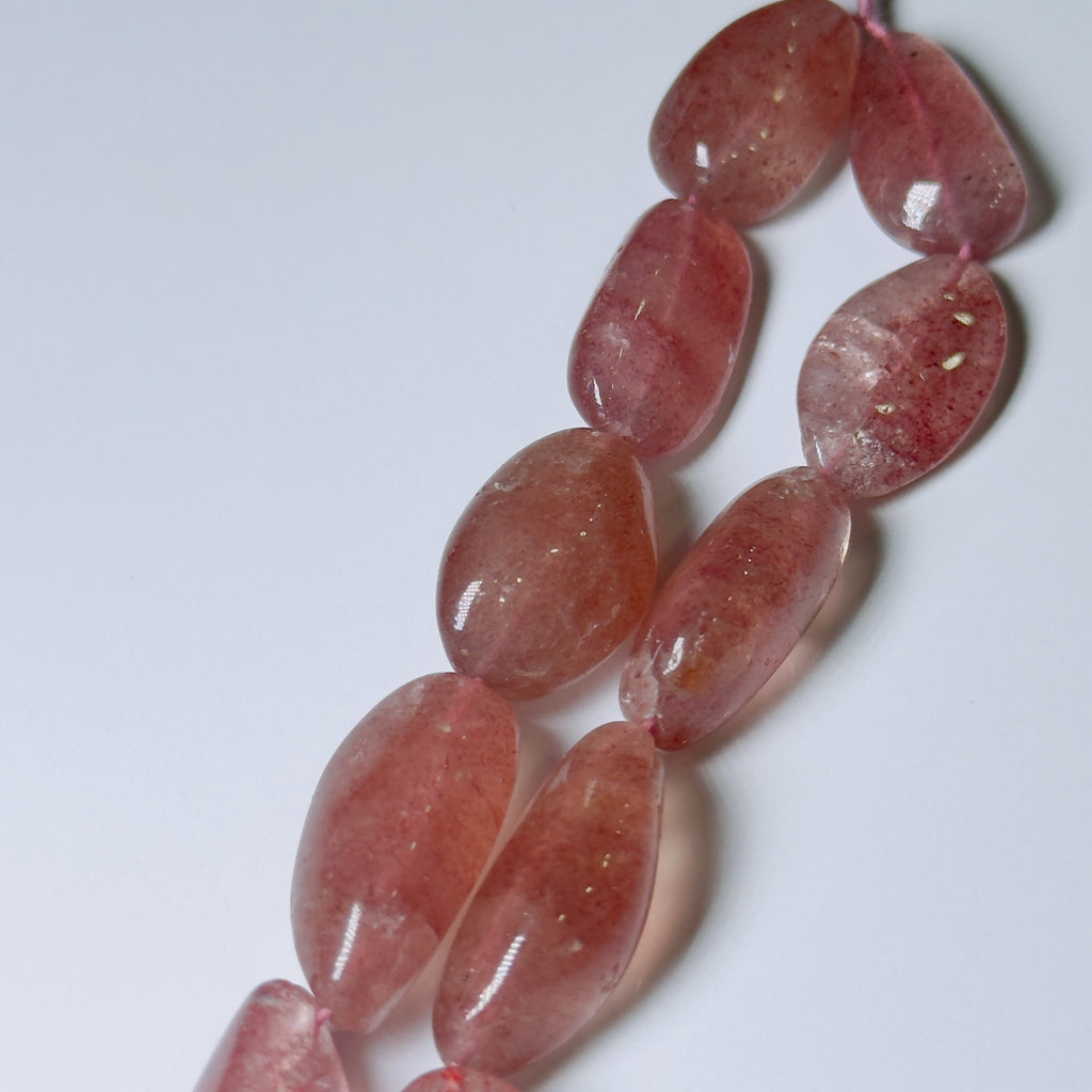 A close-up of polished oval beads in a translucent pinkish-red tone, arranged diagonally on a plain light background.