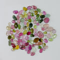 A pile of small, polished, faceted gemstones in various colors including pink, green, yellow, and purple, arranged in a loose circle on a plain light gray background.