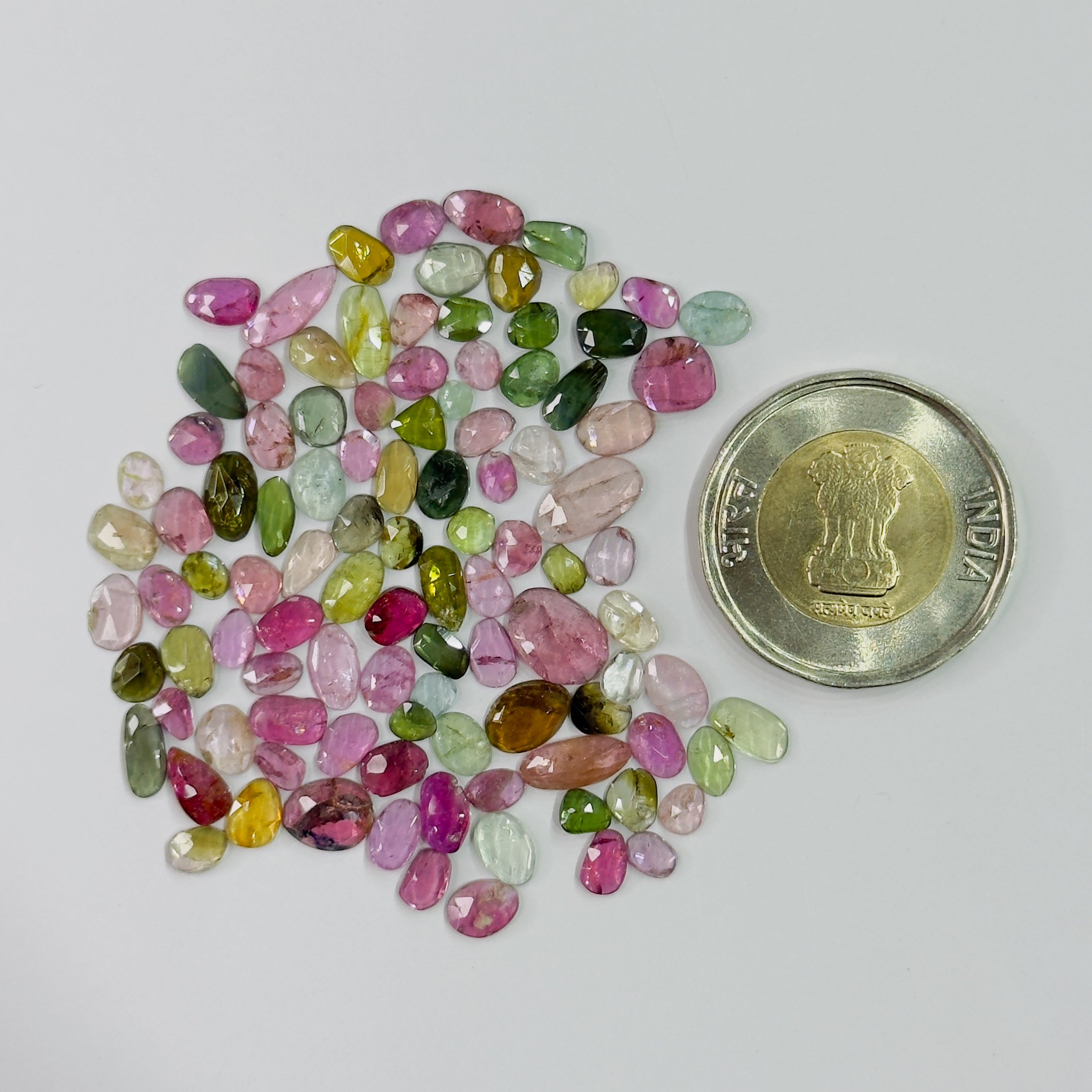 A pile of small, polished, faceted gemstones in various colors including pink, green, yellow, and purple, displayed next to a gold-colored Indian coin for scale on a plain light gray background.