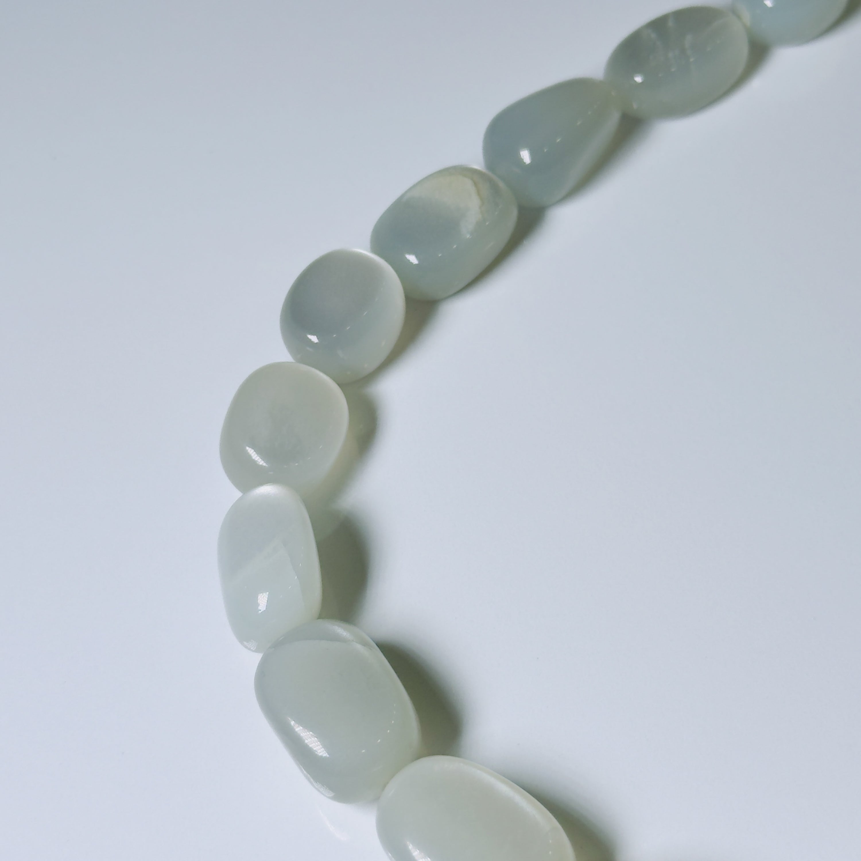 A close-up of a curved strand of polished, smooth tumbled stone beads in a pale icy blue-green hue on a clean white background.